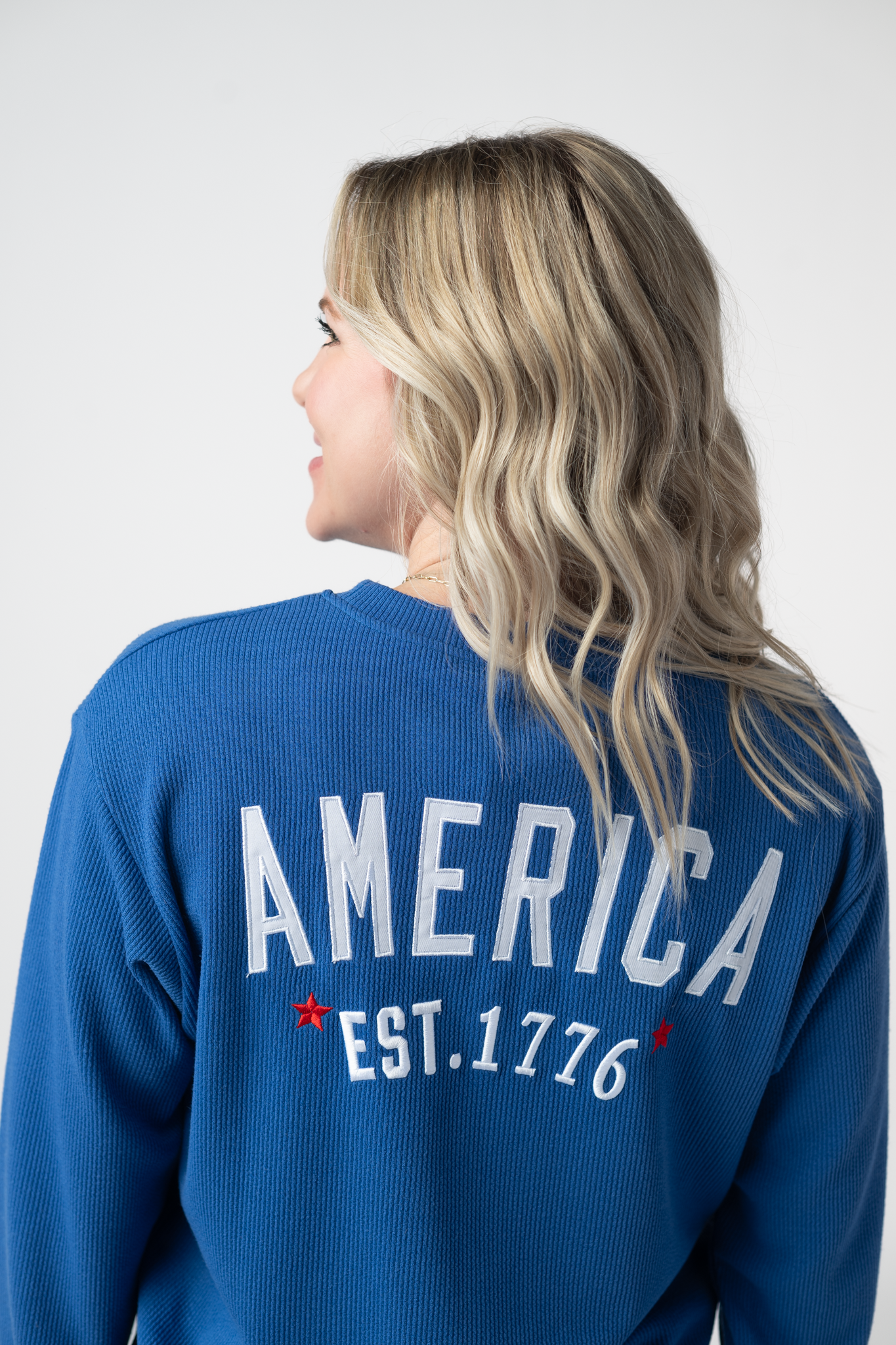 Woman wearing a blue America pullover sweatshirt with white embroidered America Est. 1776 text and red stars on the back, paired with light wash jeans