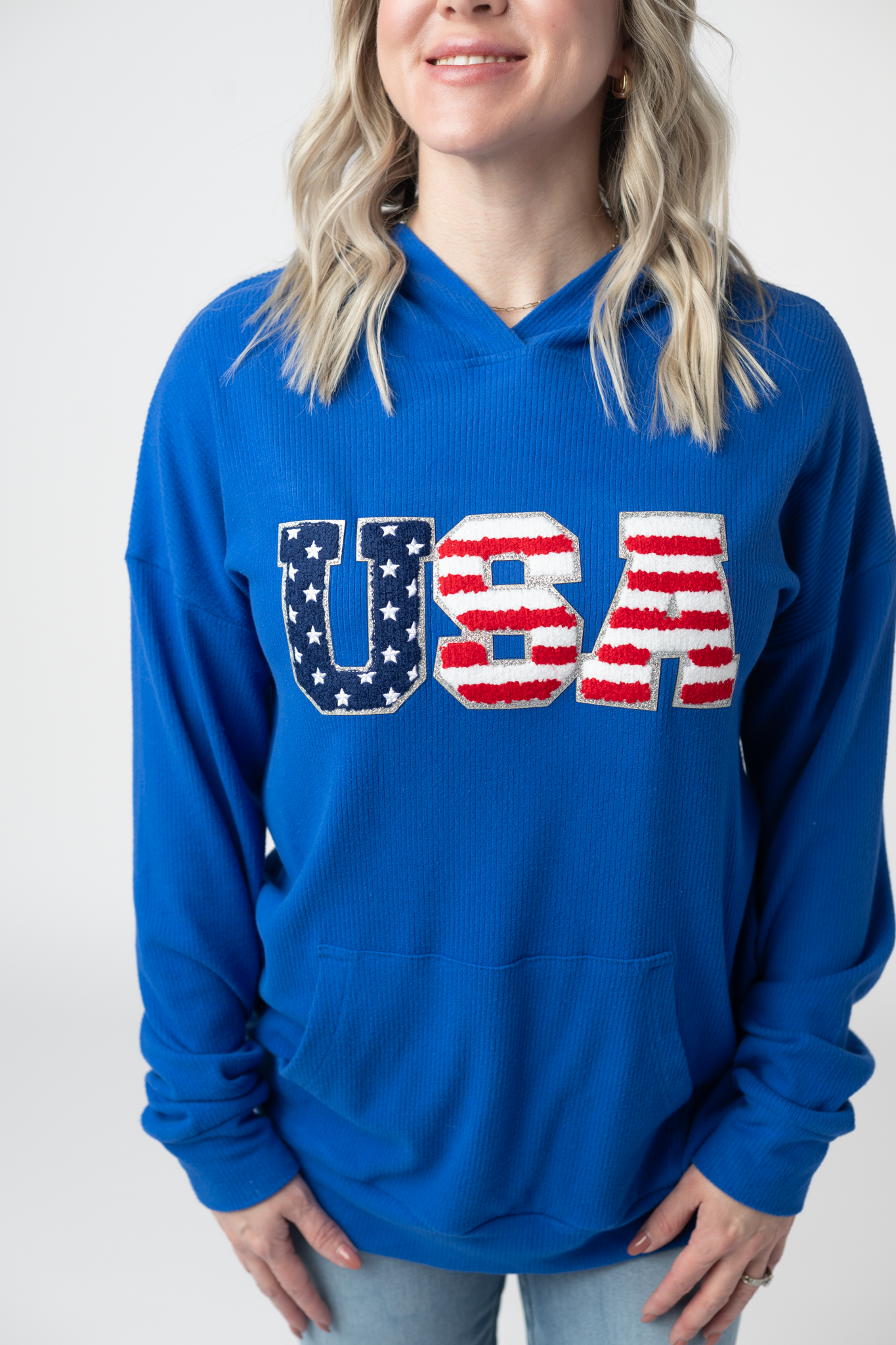 Woman wearing a royal blue hooded pullover with chenille USA patch lettering in stars and stripes design, featuring kangaroo pocket