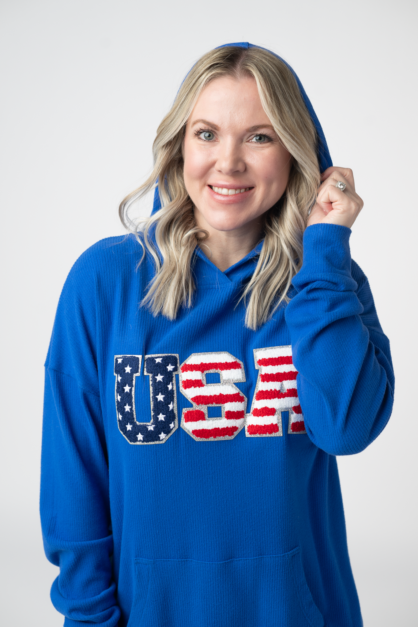 Woman wearing a royal blue hooded pullover with chenille USA patch lettering in stars and stripes design, featuring kangaroo pocket