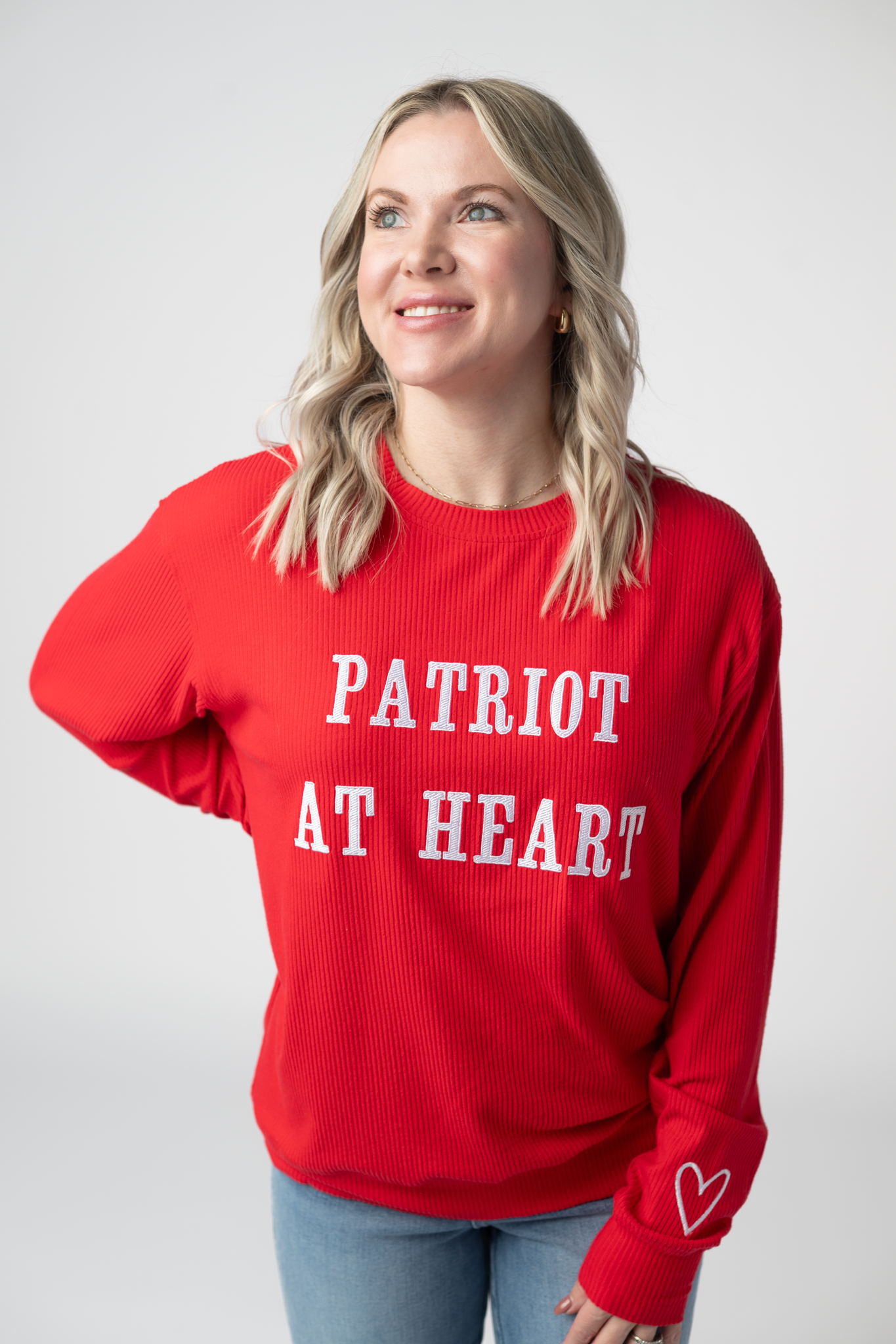 Woman wearing a red ribbed crew neck pullover sweater with white embroidered Patriot at Heart text and heart detail on sleeve cuff, paired with light wash jeans