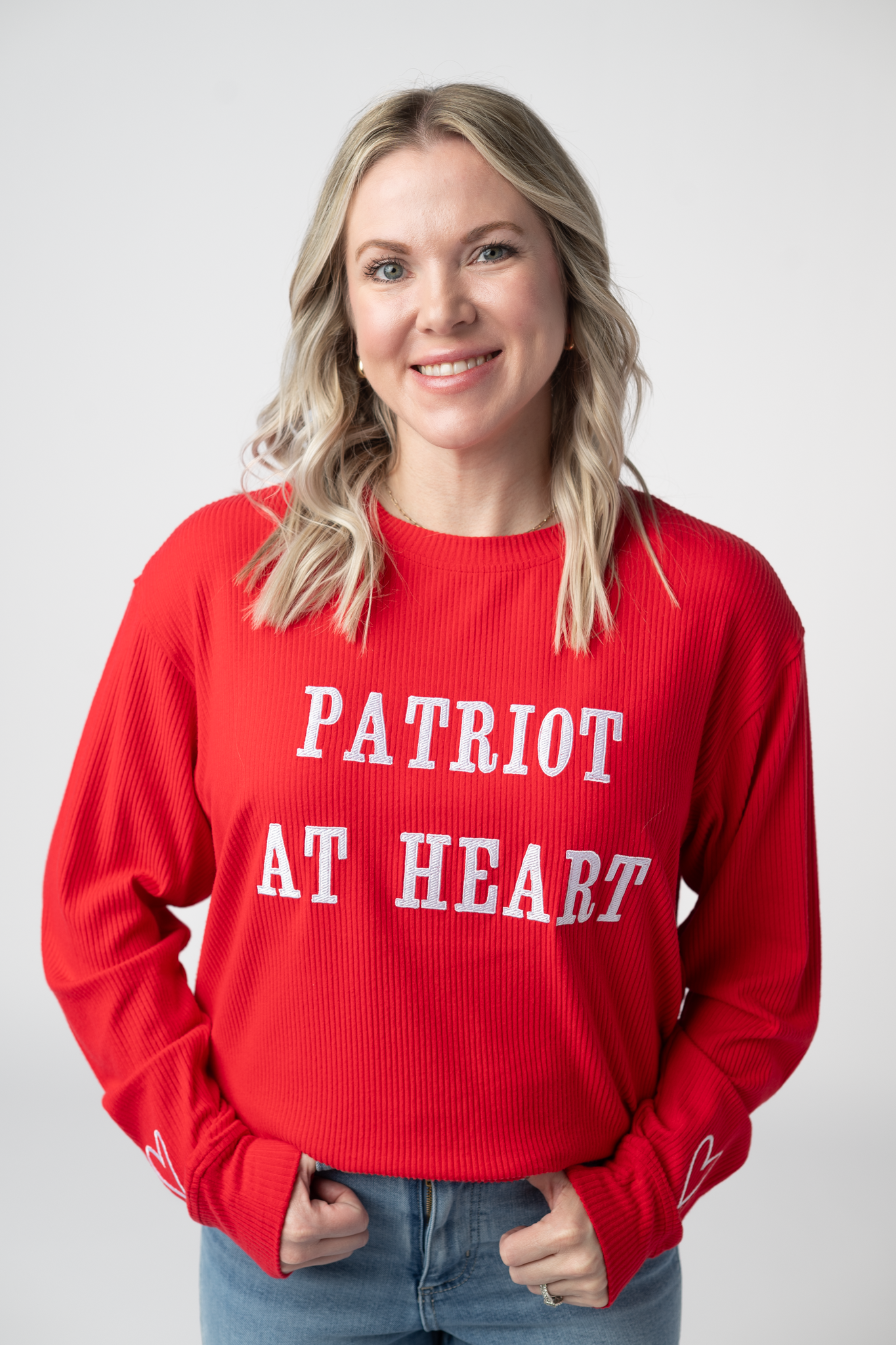 Woman wearing a red ribbed crew neck pullover sweater with white embroidered Patriot at Heart text and heart detail on sleeve cuff, paired with light wash jeans