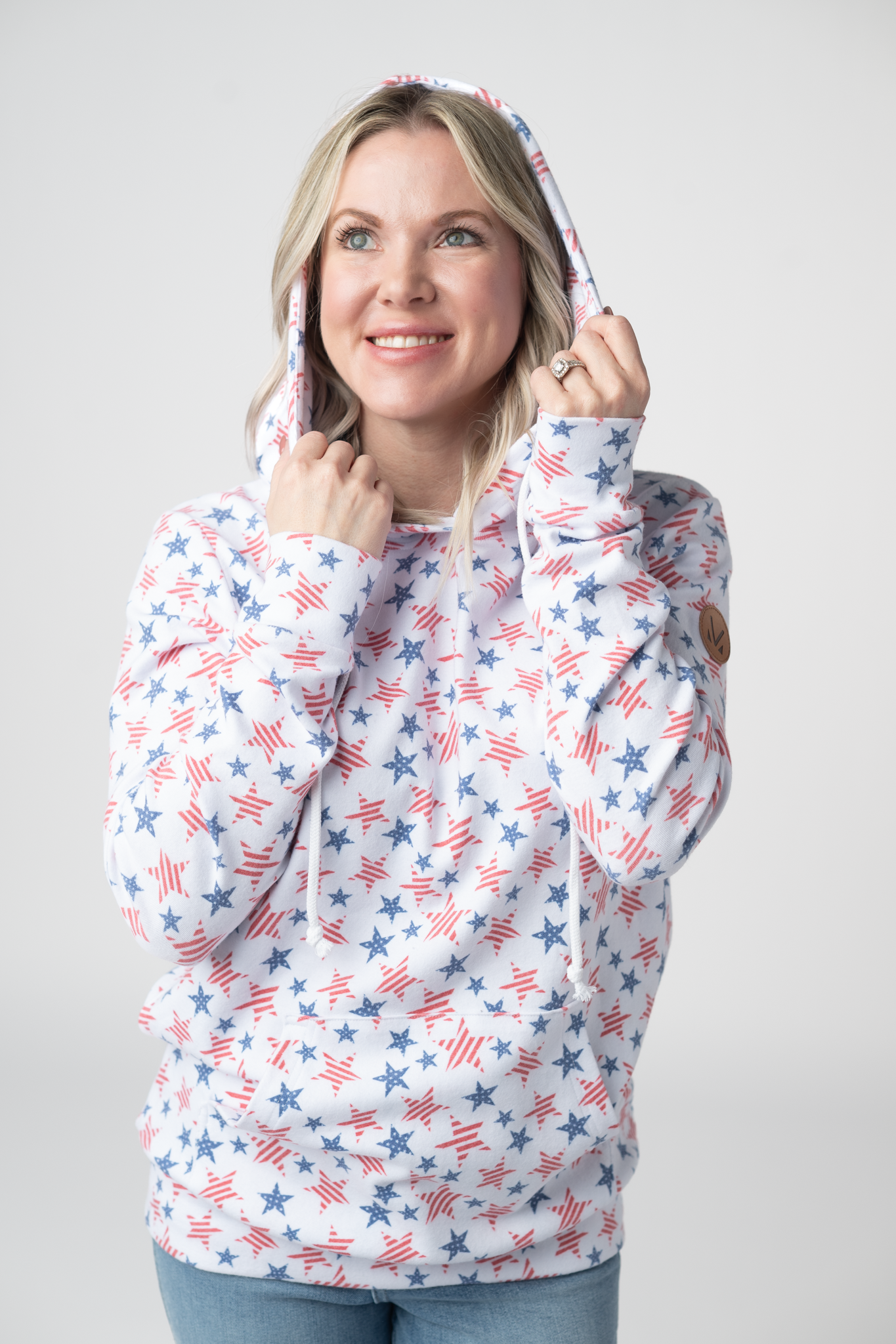 Woman wearing a white pullover hoodie with all-over red striped stars and blue solid stars print, featuring kangaroo pocket, drawstring hood, and leather logo patch