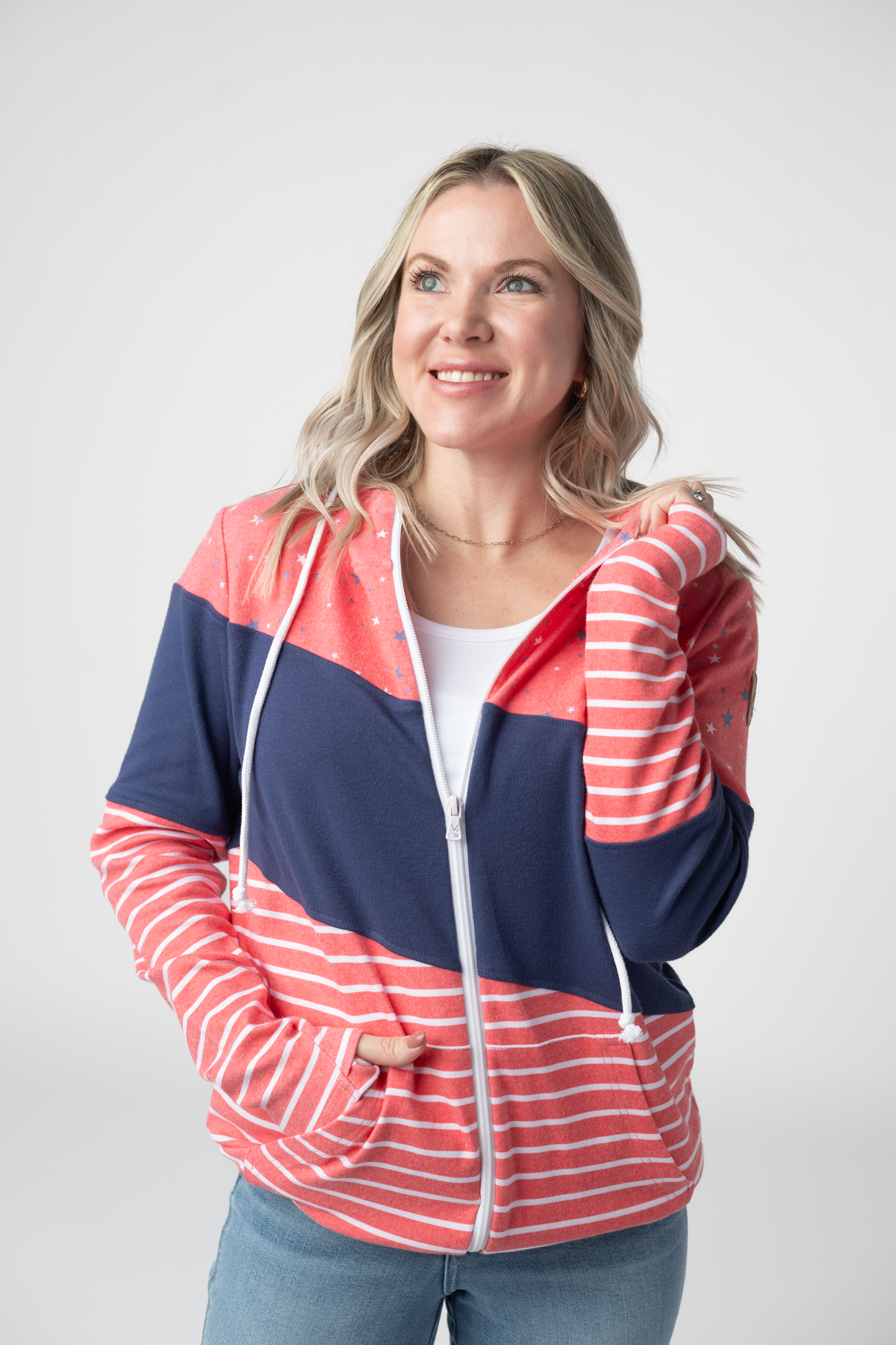 Woman wearing a patriotic colorblock full zip hoodie with red and white stripes, navy blue panels, and star print hood with white drawstrings