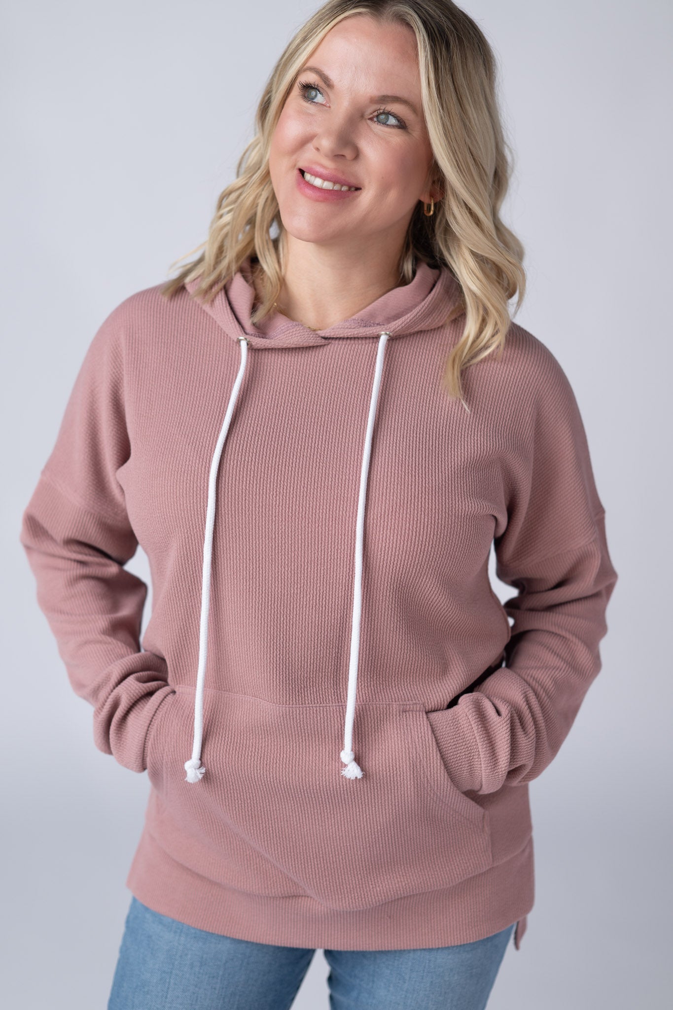 Woman wearing a dusty rose Kacey Ribbed Hoodie with white drawstrings, kangaroo pocket, and ribbed texture, paired with jeans