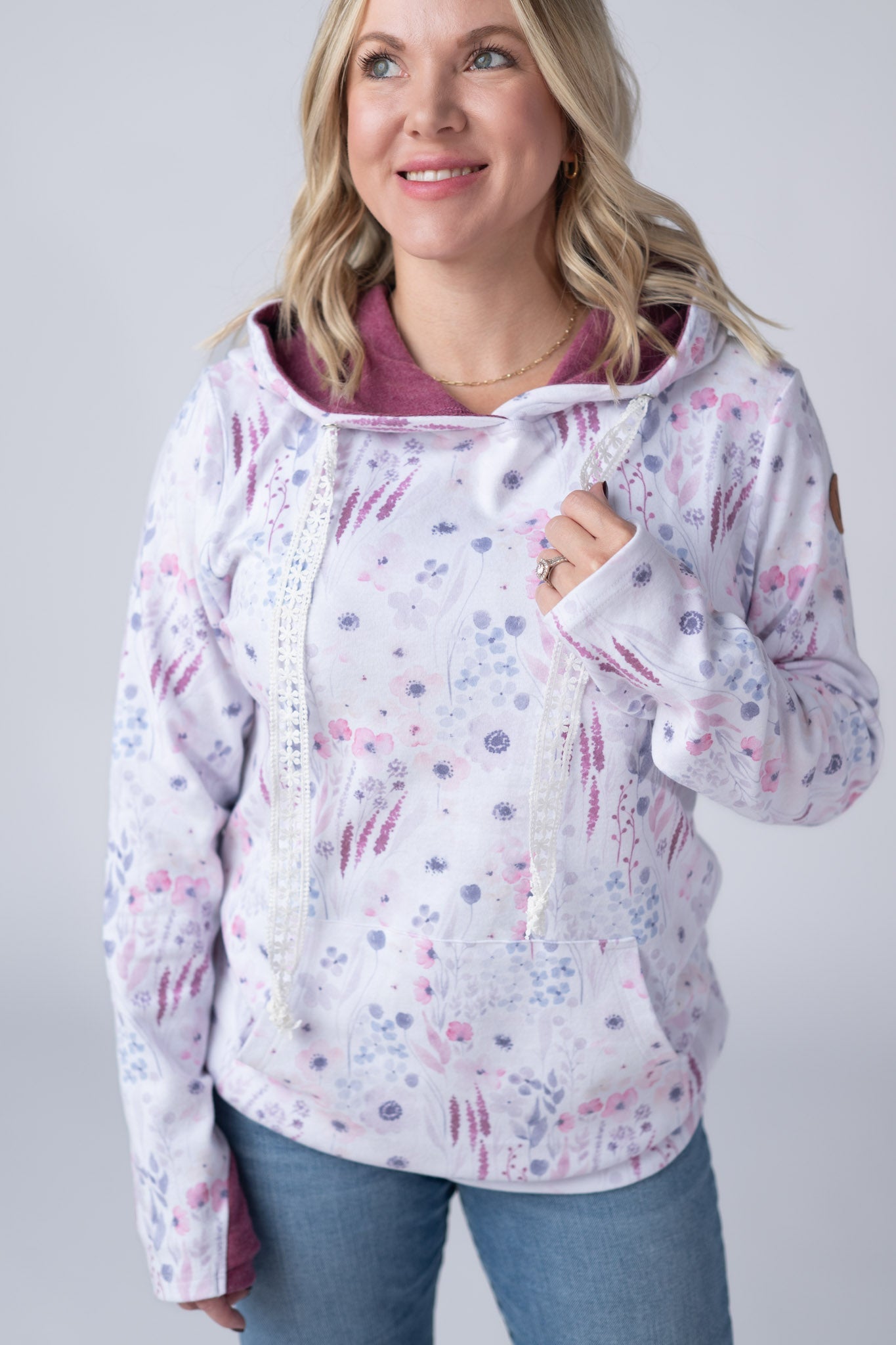 Woman wearing a white Hailey Pullover Hoodie with soft watercolor Wildflower Gardens print in pink, purple, and magenta, featuring crochet drawstrings, kangaroo pocket, and contrast mauve hood lining