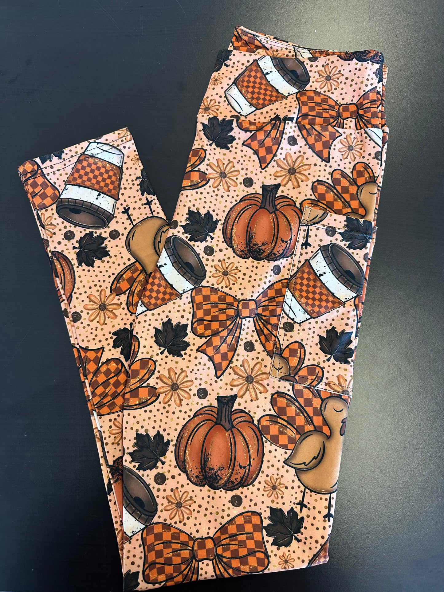 
Flat lay of peach full-length leggings with Thanksgiving print featuring pumpkins, checkered bows, coffee cups, turkeys, maple leaves, and daisies