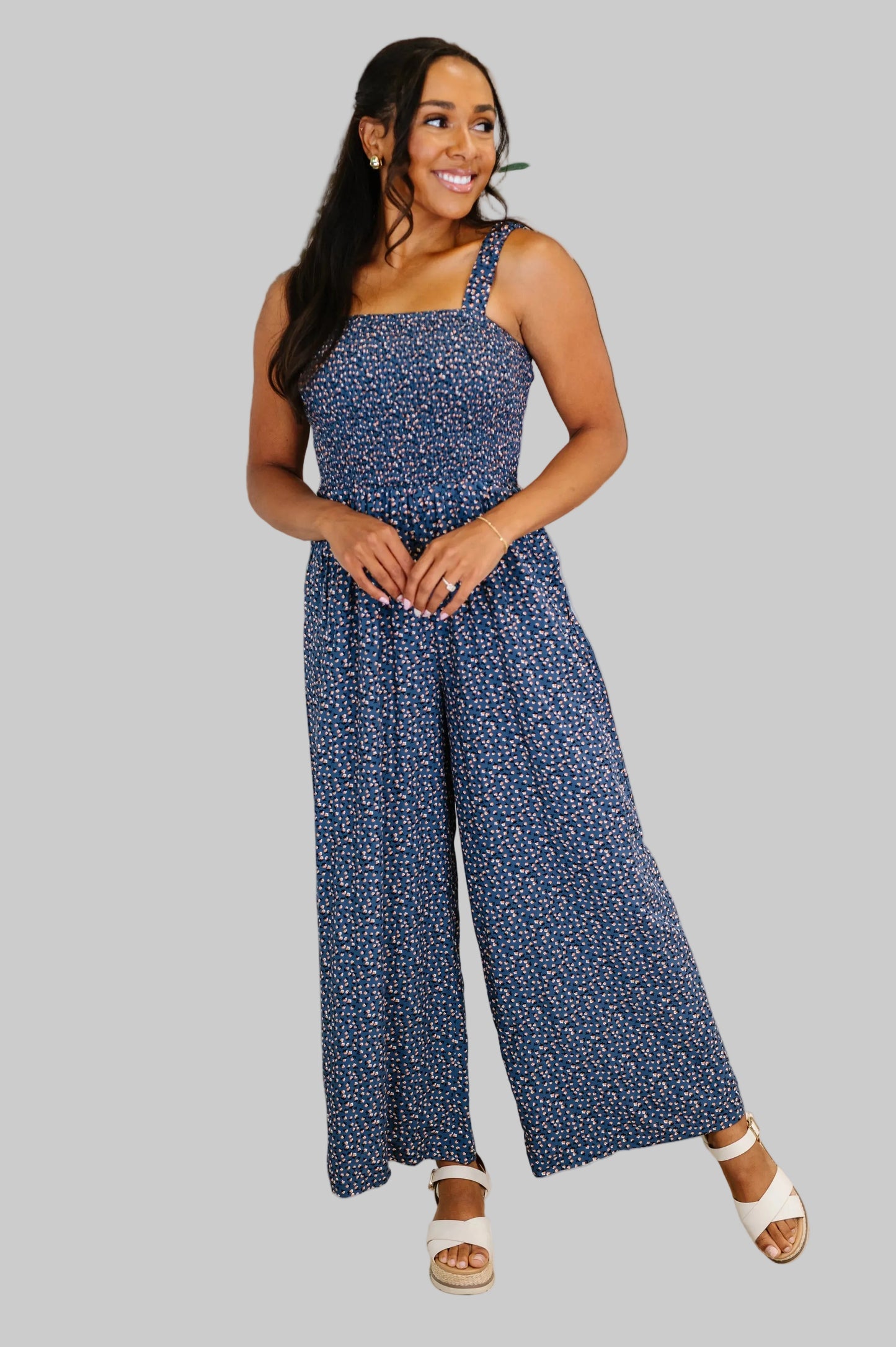 Woman wearing the Floral Wide Leg Jumpsuit in Blue Floral, a navy ditsy floral wide leg jumpsuit with smocked bodice, square neckline, elastic straps, and functional pockets, styled with white wedge sandals.