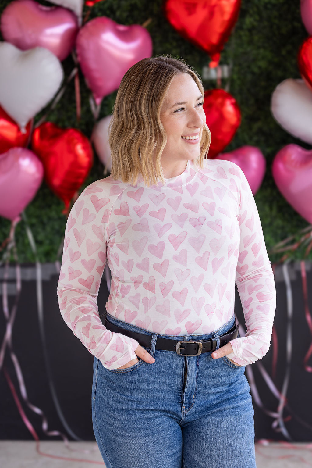 Woman wearing fitted long sleeve top with pink watercolor hearts print, high crew neckline, and thumbholes