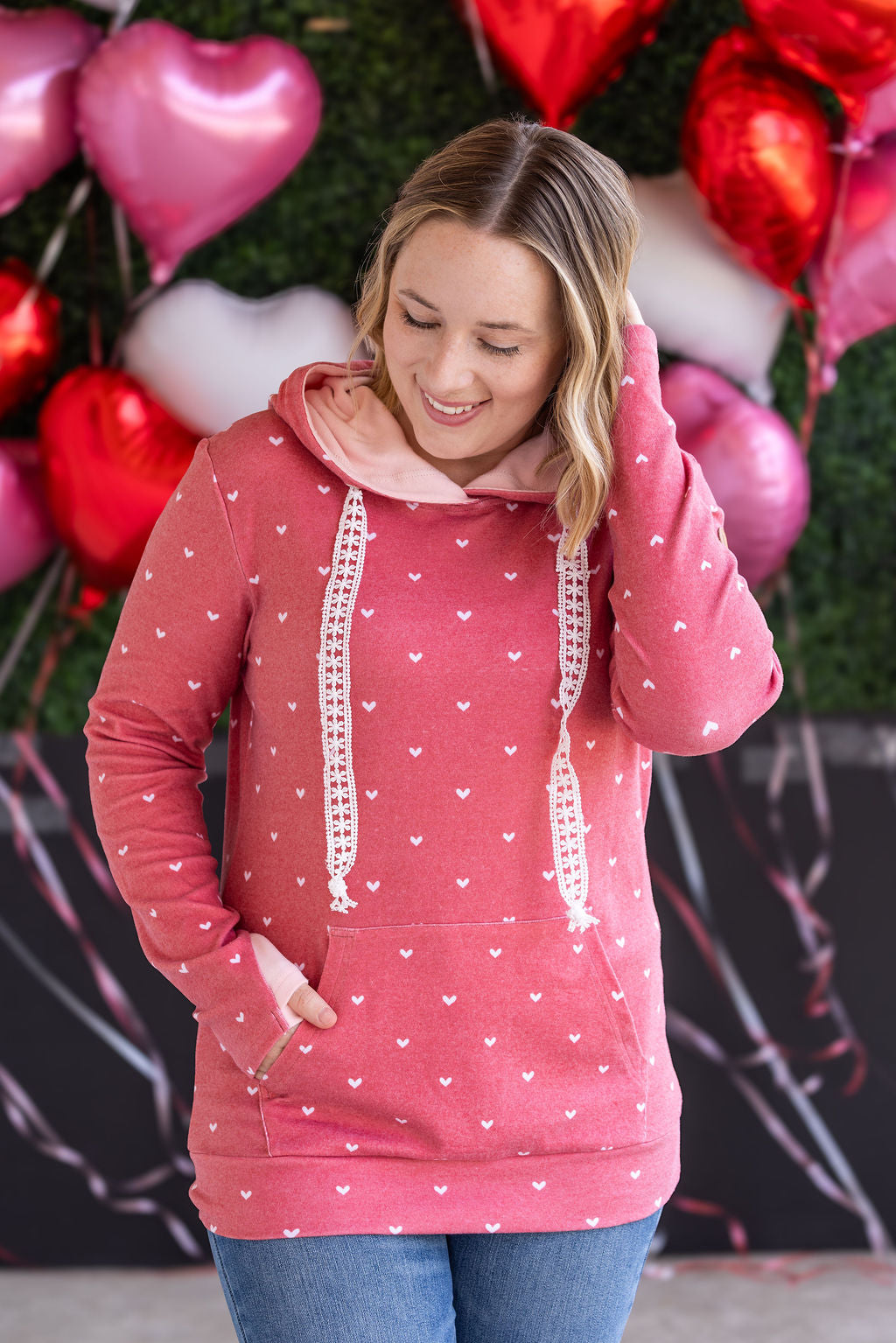 Woman wearing red pullover hoodie with white hearts print all over, featuring pink hood lining, crochet drawstrings, and kangaroo pocket
