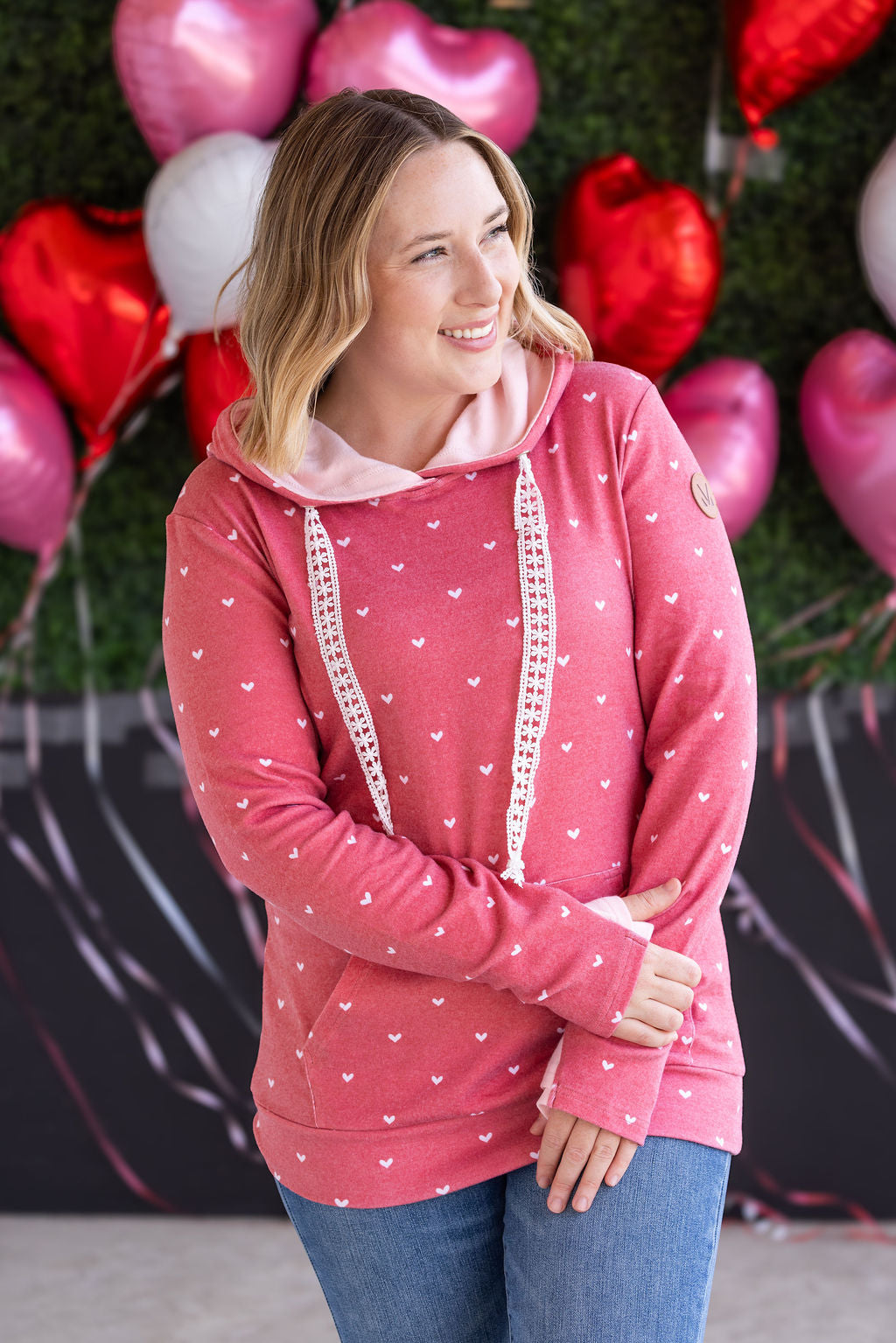 Woman wearing red pullover hoodie with white hearts print all over, featuring pink hood lining, crochet drawstrings, and kangaroo pocket