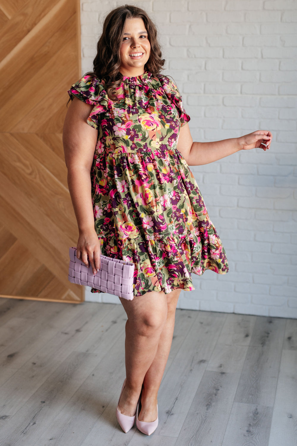 A woman with long, wavy brown hair stands indoors, wearing a floral dress with vibrant pink, green, and yellow patterns. She is smiling and holding a light purple woven clutch. The dress features ruffled short sleeves and a tiered skirt. She completes the outfit with light purple heels. The background includes a white brick wall and a wooden panel.