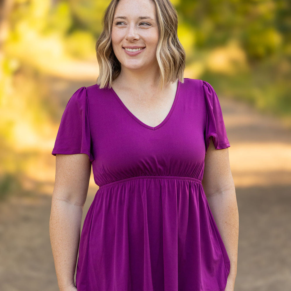 A woman with shoulder-length blonde hair stands outdoors, wearing a  purple colored V-neck top. She is smiling, with a blurred background of greenery and sunlight. The top features short sleeves and is fitted at the bust, flowing out slightly below.