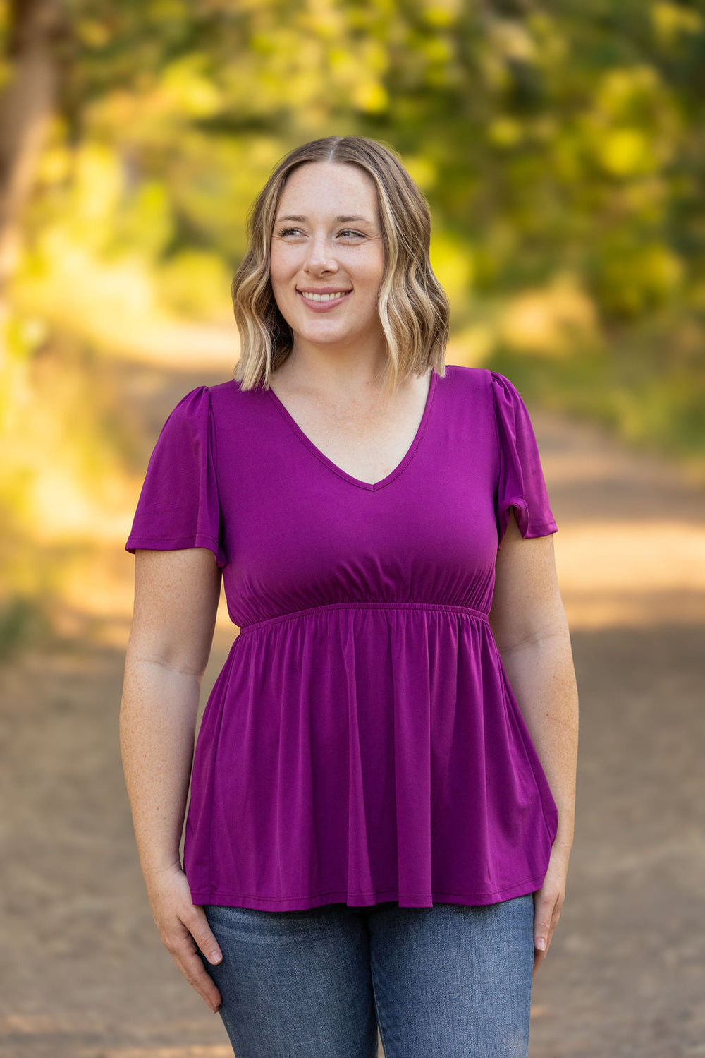 A woman with shoulder-length blonde hair stands outdoors, wearing a  purple colored V-neck top. She is smiling, with a blurred background of greenery and sunlight. The top features short sleeves and is fitted at the bust, flowing out slightly below.