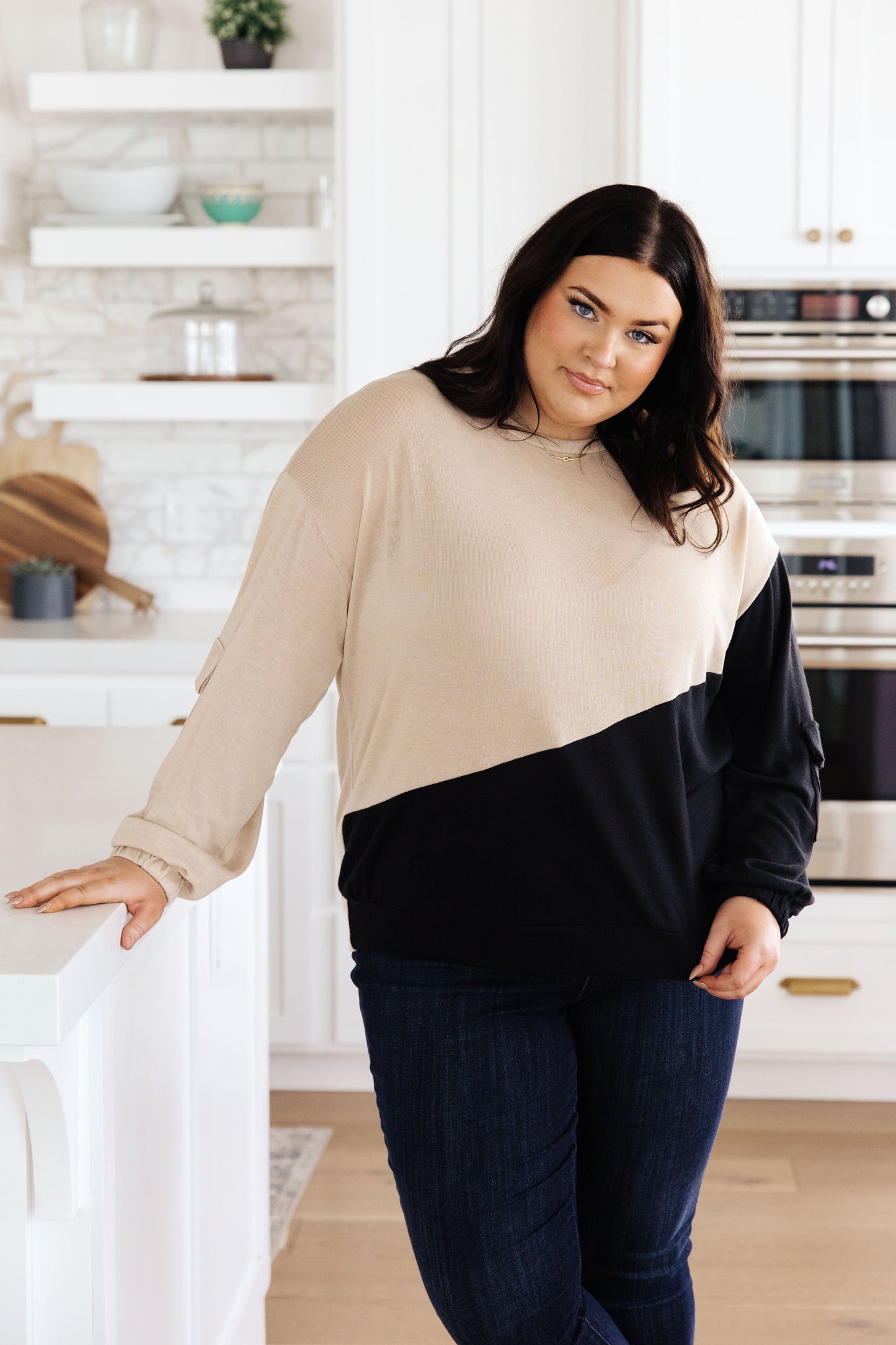Woman wearing a taupe and black asymmetrical colorblock long sleeve top with dropped shoulders and cargo sleeve pocket, paired with dark jeans