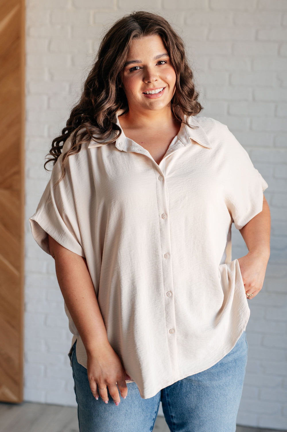 A woman with long, wavy brown hair stands indoors, wearing a light beige button-up shirt with rolled-up sleeves. She is smiling and has one hand in her pocket. The background features a white brick wall and a wooden panel.