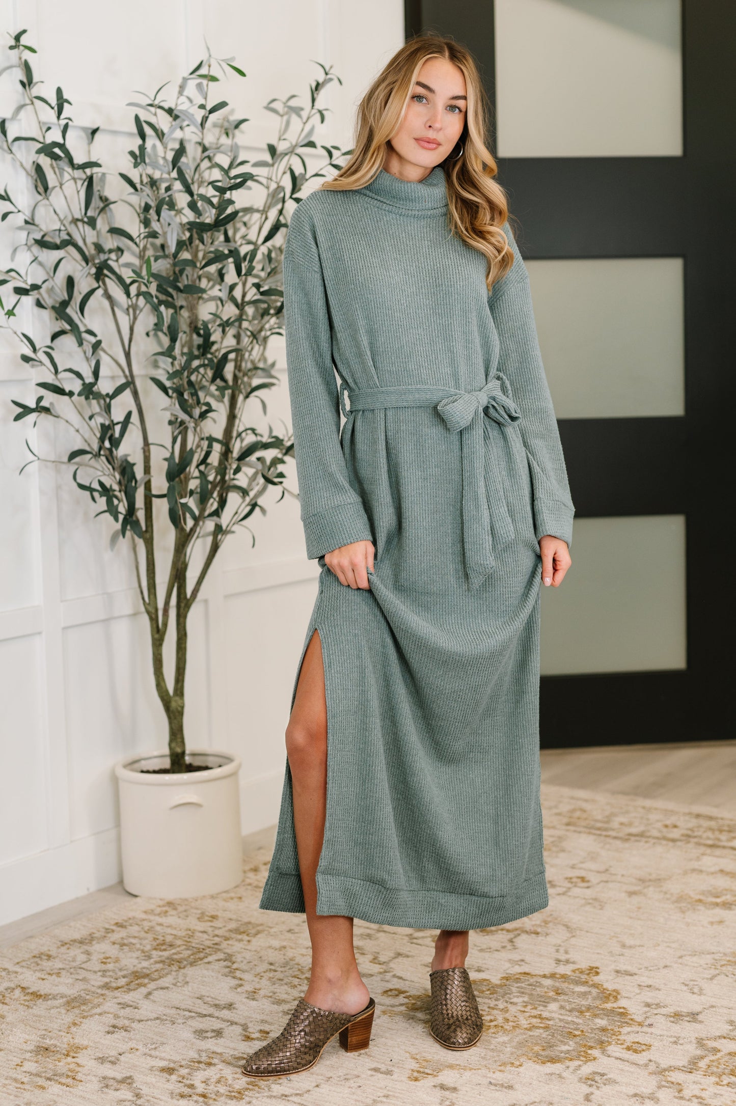 Woman wearing a soft teal waffle knit maxi dress with turtleneck, long sleeves, and waist tie belt, paired with woven mules