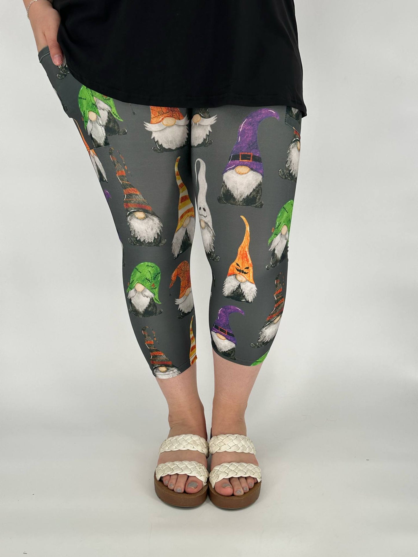 Model wearing gray capri leggings with Halloween gnome print featuring witch, ghost, and jack-o-lantern gnomes, with square side pockets, styled with a black top and cream sandals