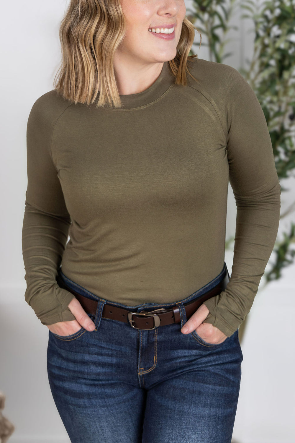 Woman wearing an olive green long-sleeve top and blue jeans with a blurred background