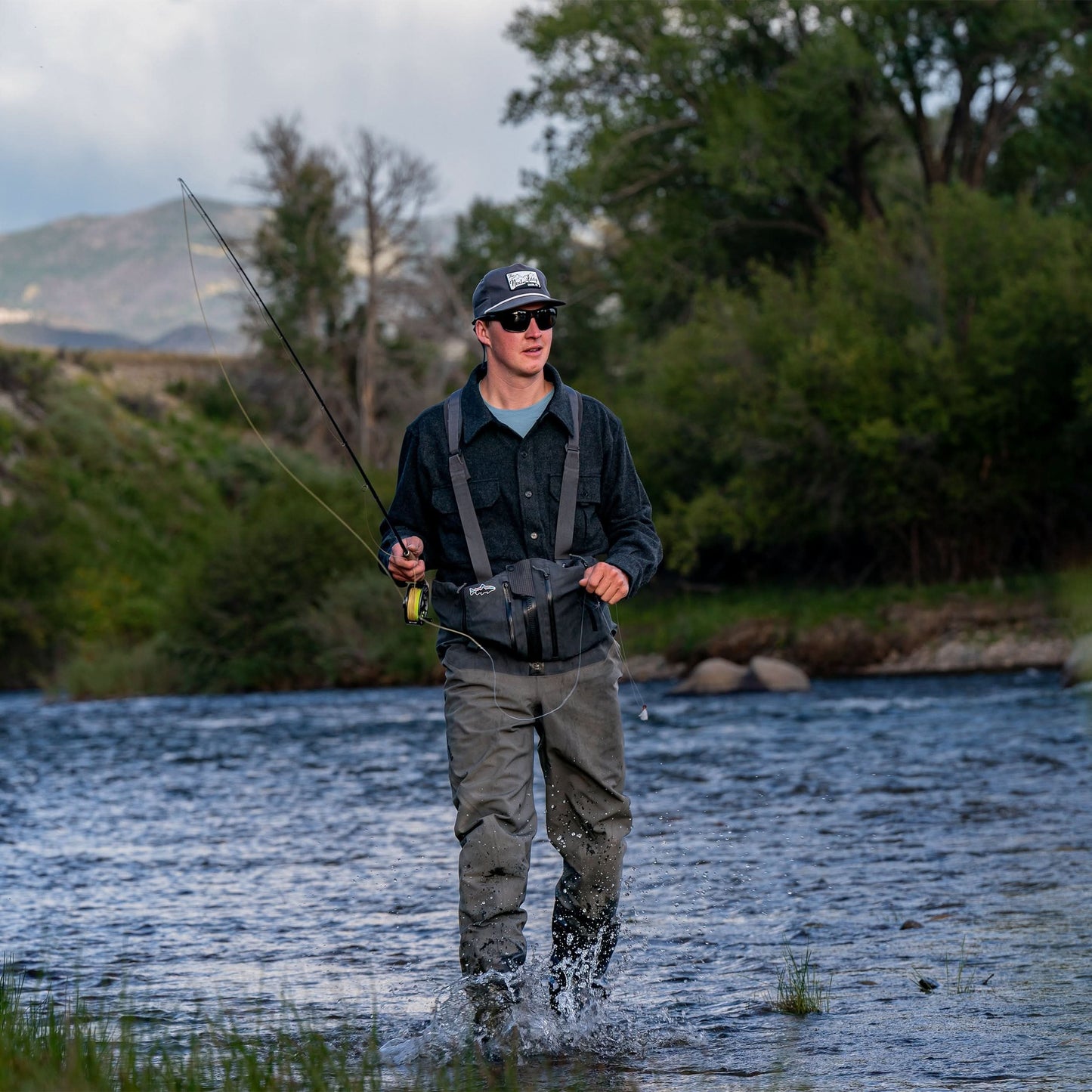 Man fishing in a river with mountains and trees in the background