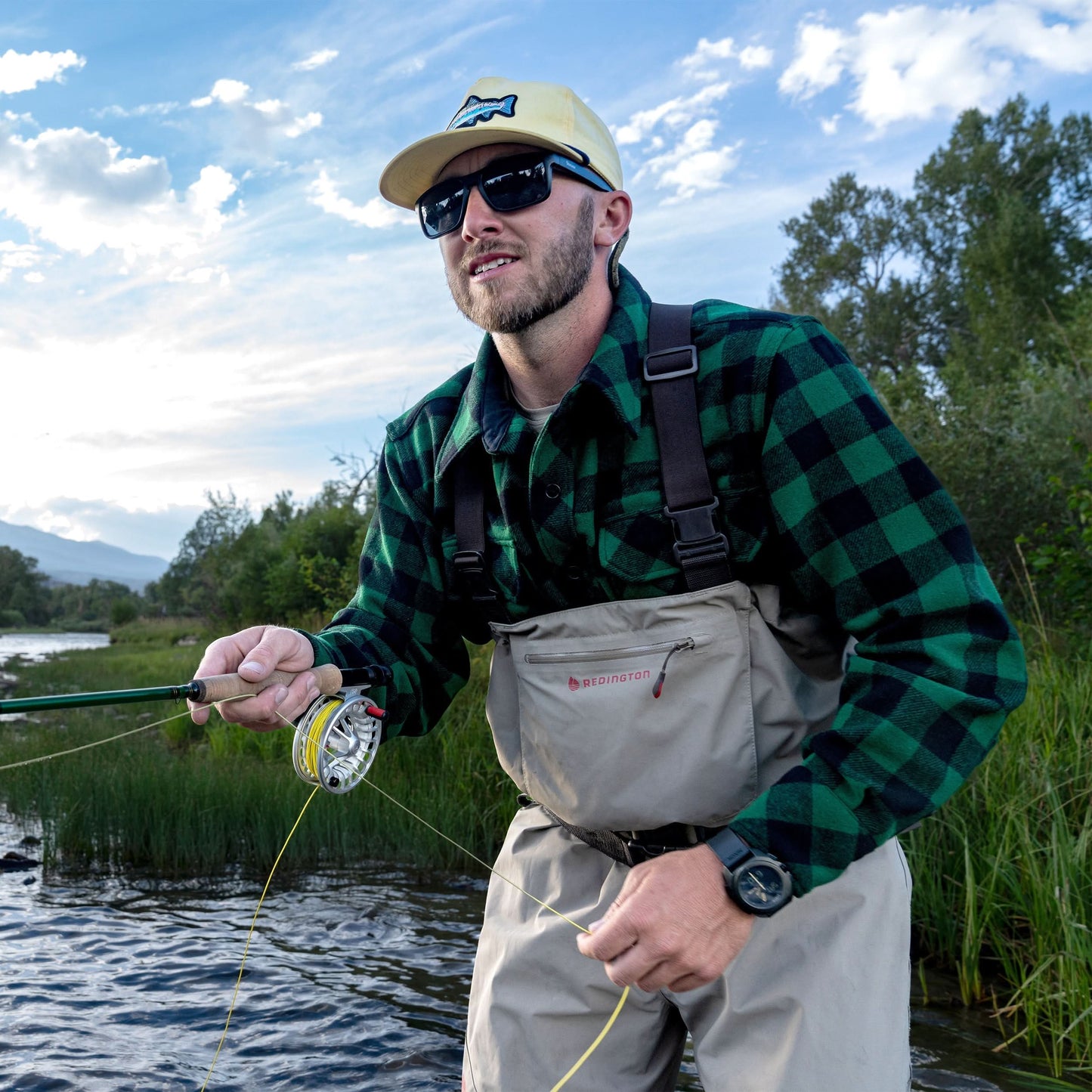 Man fishing by a lake with a green checkered shirt and beige waders.