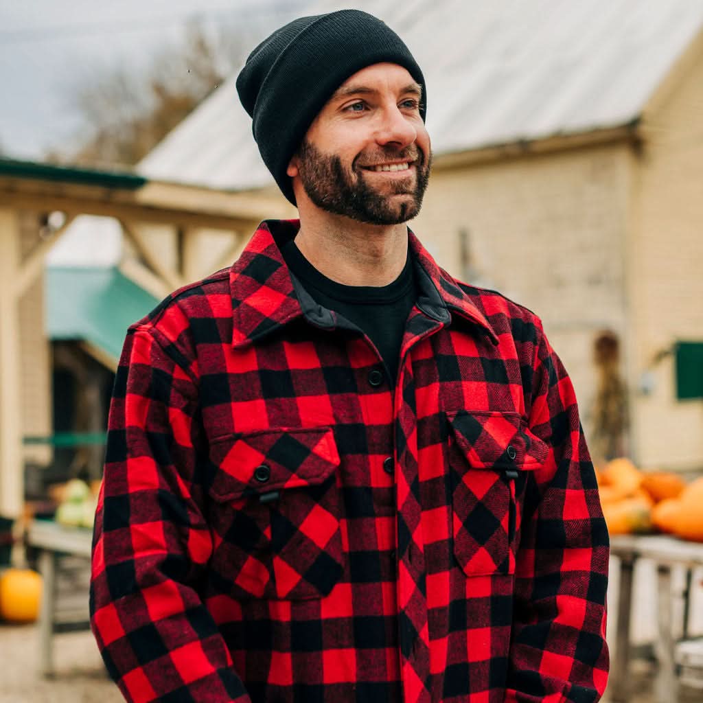Man wearing a red and black plaid shirt and black beanie outdoors with pumpkins in the background
